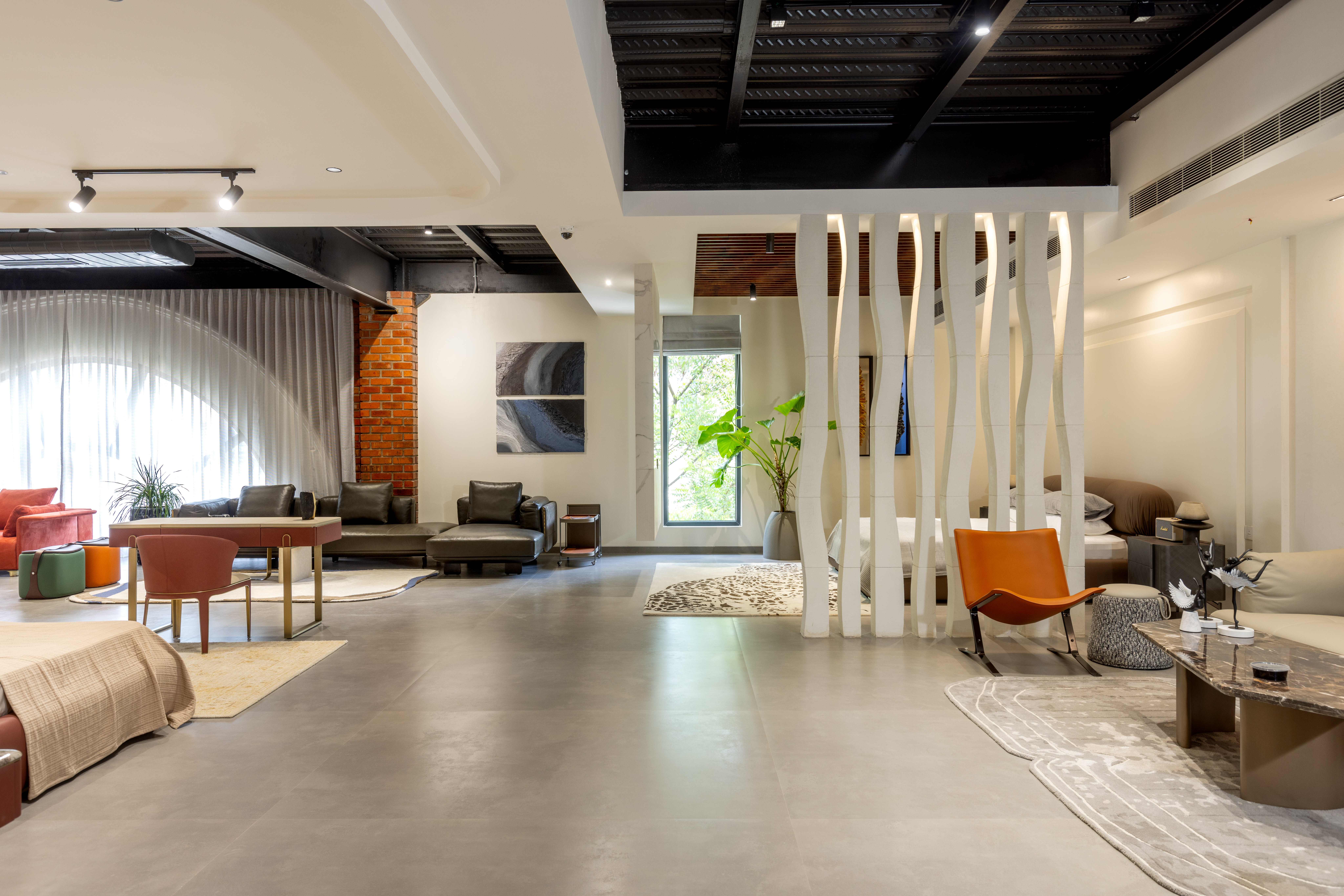 Double-height showroom hall with separate seating territories: a black leather sofa on a navy geometric rug, a navy sofa on an amber rug, a plum recliner, and a dark face-shaped vase on a plinth beneath tall sheer curtains.
